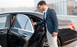 Chauffeur opens the rear door of a black sedan with a smile, greeting a passenger—reflecting punctual, professional service.