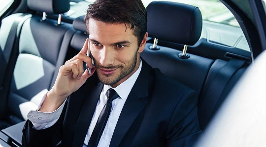 Confident businessman in a sharp suit sits in the backseat of a luxury car, speaking on the phone with a focused smile—capturing a moment of professional travel and productivity on the go.