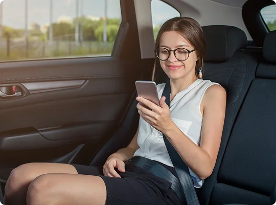 A young woman wearing glasses relaxes in the backseat of a car, smiling as she looks at her phone—capturing a moment of comfort and enjoyment during a smooth ride.