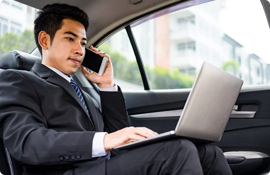Young professional in a suit sits in the backseat of a car, multitasking with a laptop on his lap and a phone to his ear—capturing productivity and focus during executive travel.