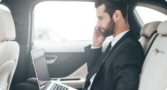 Businessman in a suit sits in the backseat of a luxury car, speaking on the phone and working on a laptop—capturing a moment of productivity and professionalism during travel.