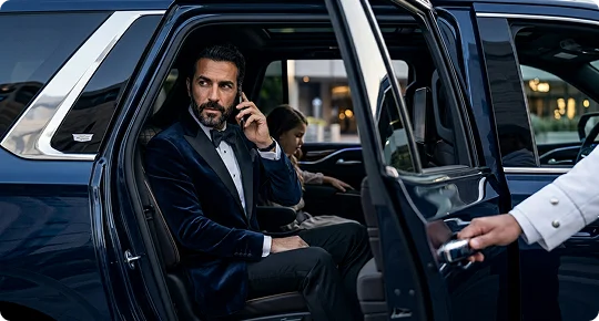 A man wearing a blue velvet tuxedo jacket and bowtie speaks on a smartphone in the back seat of a luxury vehicle. An open door is held by a driver wearing a white shirt cuff, with a young girl sitting further back in the cabin.